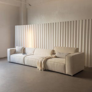 May include: A long, cream-colored modular sofa featuring a low-profile design with wide armrests, a rectangular backrest, and thick seat cushions. The sofa is accented with a square throw pillow, a rectangular lumbar pillow, and a draped fringe throw.
