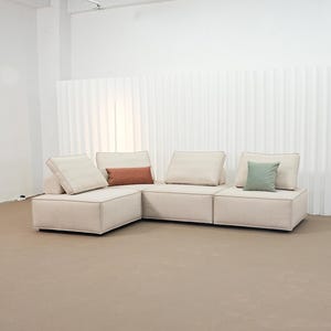 May include: A modular sectional sofa upholstered in light beige fabric with visible piping seams. The sofa consists of three connected square base sections and four back cushions, accented by one rectangular rust-colored pillow and one square sage green pillow.