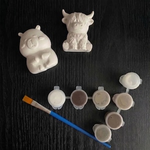 May include: Two unpainted white ceramic figurines, one shaped like a bear and one like a Highland cow, accompanied by a set of six small connected paint pots in neutral tones and a single paintbrush with a blue handle and a flat orange tip.