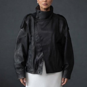 May include: A black leather jacket featuring a high collar with double button closures, an asymmetrical front overlap, structured shoulders with epaulets, and long sleeves with cuffs. The jacket is worn over a plain white garment.