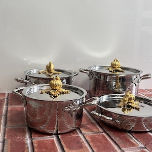 Puede incluir: Un juego de cuatro ollas y sartenes de metal plateado con textura martillada. Cada tapa presenta un asa dorada en forma de calabaza con hojas. El conjunto incluye dos ollas profundas con asas laterales y dos sartenes más bajas a juego.