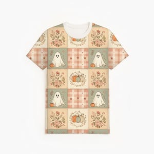 May include: A short-sleeve t-shirt featuring a repeating patchwork pattern of muted autumn colors. The design includes squares with white ghosts holding orange pumpkins, floral arrangements, and plaid squares with small stars in soft peach and sage green.