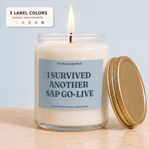 May include: A clear glass jar candle with a lit wick and a gold-colored metal lid. The label is light blue with dark text reading TheMoonLightWick, I SURVIVED ANOTHER SAP GO-LIVE, and %100 SOY WAX CANDLE. MADE IN USA. A small sign above shows 5 label colors.