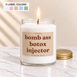 May include: A white soy wax candle in a clear glass jar with a lit wick. The label is tan with brown text that reads "bomb ass botox injector" and "TheMoonlightWick". A gold metal lid sits beside the jar. A small graphic shows 5 label colors.
