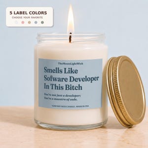 May include: A white soy wax candle in a clear glass jar with a gold lid. The light blue label features the text: TheMoonLightWick, Smells Like Software Developer In This Bitch, You're not just a developer; You're a maestro of code. 100% SOY WAX CANDLE.