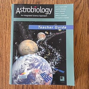 Puede incluir: Un libro de texto titulado Astrobiology: An Integrated Science Approach, Teacher Guide. La portada presenta una escena espacial con la Tierra, un planeta, un satélite y estrellas sobre un fondo oscuro. Los autores son Asbell-Clarke, Barstow y otros.