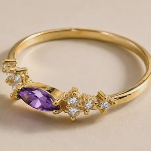 May include: A gold ring featuring a central marquise-cut purple amethyst gemstone flanked by two clusters of three small round clear white stones on each side, set along a thin polished gold band with a smooth and reflective metallic finish.