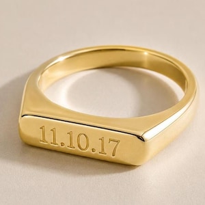 May include: A polished gold-tone signet ring featuring a flat rectangular face engraved with the date 11.10.17. The ring has a smooth, rounded band and a high-shine metallic finish, showcasing a clean, solid construction with clear lettering.