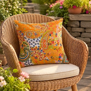 Könnte beinhalten: Ein quadratisches Zierkissen mit leuchtend orangefarbenem Hintergrund und einem mehrfarbigen Paisley- und Blumenmuster in Blau, Rot, Gelb und Grün. Das Kissen liegt auf einem hellbraunen Korbstuhl mit einem beigen Sitzpolster.