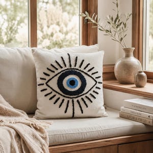 May include: A square off-white throw pillow featuring a centered, tufted evil eye design. The eye is composed of a central light blue pupil, a dark blue iris, and a black outline with radiating black lashes embroidered on the fabric surface.
