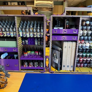 May include: A large, purple, modular storage case with two open sides, featuring numerous shelves and drawers filled with various paint bottles, small pots, and art supplies. The interior includes organised racks for dropper bottles and stacked paint pots.