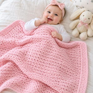 May include: A soft, light pink crocheted baby blanket with a textured, grid-like stitch pattern. The blanket features a solid, uniform weave throughout, providing a consistent look and feel for a nursery setting or as a gift for a newborn.