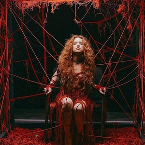 May include: A person with long curly red hair sits in a chair, wearing a red dress with long fringe details. The person is surrounded by a complex web of red yarn or string that extends from the chair to the surrounding dark frame structure.