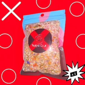 May include: A clear plastic resealable pouch filled with a mixture of pink salt crystals and dried botanical pieces. A circular black label on the front features a red cross design, a rose emblem, and the white text Foxy's Co.X.