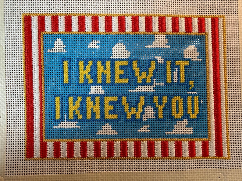May include: A needlepoint canvas featuring a blue sky background with white clouds and the yellow text I KNEW IT, I KNEW YOU. The design is framed by a border of vertical red and white stripes, all set against a white grid mesh background.