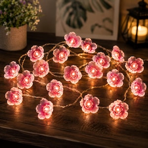 May include: A string of decorative LED lights featuring translucent pink flower-shaped covers. Each individual blossom contains a small, glowing light bulb at its center, connected by a thin, flexible wire arranged in a loose, coiled formation.