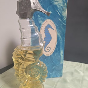May include: A clear glass perfume bottle shaped like a seahorse with a metallic silver seahorse head cap. The bottle contains a pale yellow liquid. Behind the bottle stands a rectangular blue box featuring a white oval with a blue seahorse illustration.