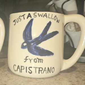 Può includere: Due tazze in ceramica color bianco sporco con un'illustrazione dipinta a mano di una rondine blu. Il testo "JUST A SWALLOW from CAPISTRANO" è scritto in nero sulla parte anteriore di ogni tazza, dotata di un manico laterale curvo.