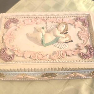 May include: A rectangular cream-colored decorative box featuring a raised relief of a cherub with wings on the lid. The lid is framed by ornate pink and mauve scrollwork. The sides of the box are detailed with gold-toned shell accents.