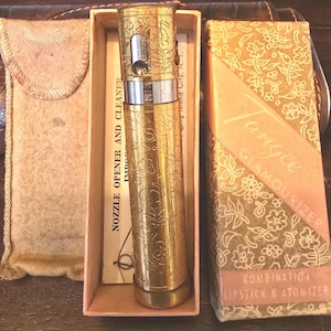 May include: A gold-toned Tangee Glamourizer combination lipstick and atomizer in a rectangular box with a floral pattern. The metal tube features an engraved flower design, a silver-colored band, and a nozzle. A tan fabric pouch is included nearby.
