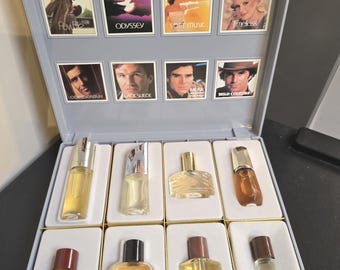 May include: A gray plastic display case containing eight miniature perfume bottles. The lid features eight square images with labels including Odyssey, Soft Musk, Timeless, Cordovan, Black Suede, Musk for Men, and Wild Country. Each bottle is clear glass.