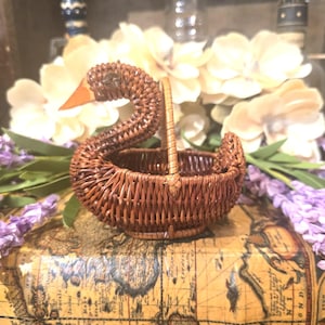 May include: A brown woven wicker basket shaped like a swan with a long curved neck, a pointed orange beak, and a small handle. The basket features a detailed woven texture throughout its body and a circular base, crafted from thin brown reeds.