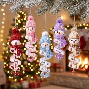 May include: Five hanging snowman ornaments with knit hats and scarves, each featuring a spiraling ribbon with a name: Keith, Mabel, Lionel, Coral, and Belen. The snowmen have unique color themes including red, pink, blue, purple, and tan.