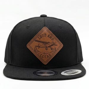 May include: A black snapback cap featuring a diamond-shaped brown leather patch on the front. The patch displays an engraved illustration of a small airplane flying over a landscape with the text LOUD AND CLEAR. A circular sticker is on the brim.