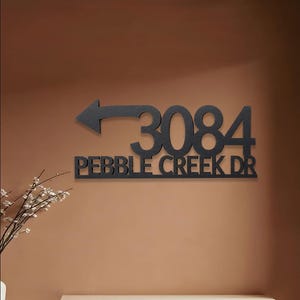 Peut inclure: Une enseigne murale en métal noir affichant le numéro de maison 3084 et le texte PEBBLE CREEK DR dans une police sans empattement en gras. L'enseigne comprend une flèche pointant vers la gauche intégrée au-dessus du nom de la rue.