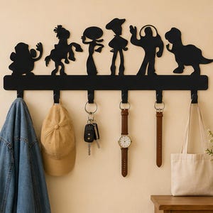 May include: A black wall-mounted coat rack featuring silhouettes of six cartoon characters along the top edge. The rack has six hooks holding a blue denim jacket, a tan baseball cap, a set of keys, a brown leather watch, a leather strap, and a tote bag.