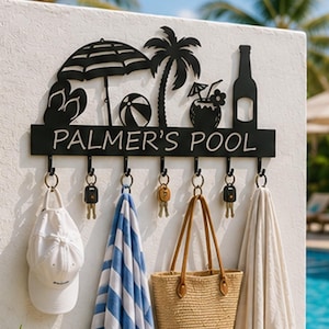 Könnte beinhalten: Eine schwarze Wandgarderobe aus Metall mit Silhouetten von Sonnenschirm, Palme, Strandball, tropischem Getränk und Flasche. Das Schild trägt den Text PALMER'S POOL und verfügt über sieben Haken mit einer weißen Kappe, Schlüsseln und Handtüchern.