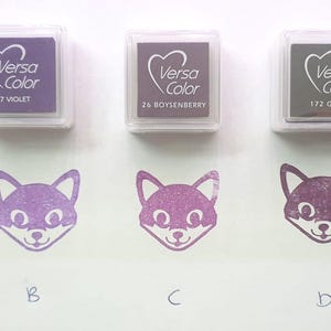 Pode incluir: Cinco almofadas de tinta de diferentes cores, etiquetadas com o nome da marca "Versa Color" e um nome de cor. As almofadas de tinta est&atilde;o dispostas em uma fila da esquerda para a direita. Abaixo das almofadas de tinta, h&aacute; cinco carimbos em forma de raposa, cada um estampado com uma tinta de cor diferente.