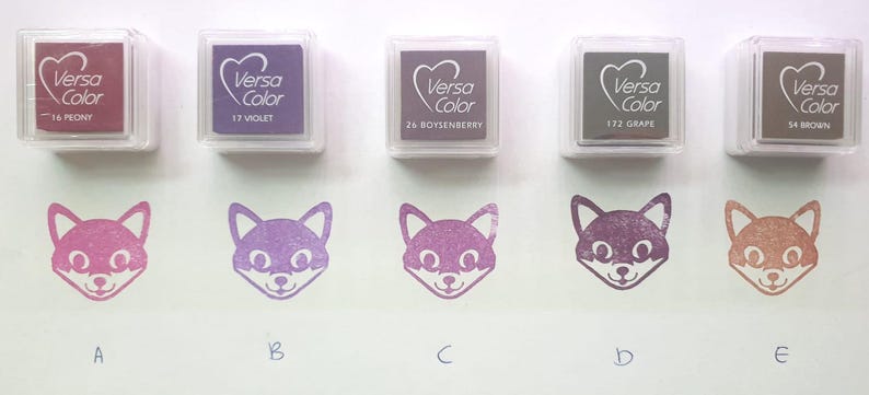 Pode incluir: Cinco almofadas de tinta de diferentes cores, etiquetadas com o nome da marca "Versa Color" e um nome de cor. As almofadas de tinta est&atilde;o dispostas em uma fila da esquerda para a direita. Abaixo das almofadas de tinta, h&aacute; cinco carimbos em forma de raposa, cada um estampado com uma tinta de cor diferente.
