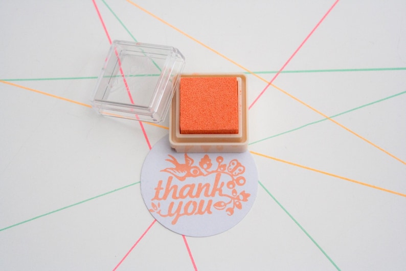 Pode incluir: Uma pequena almofada de tinta laranja em uma caixa de pl&aacute;stico transparente com um adesivo branco que diz "obrigado" com um design floral.