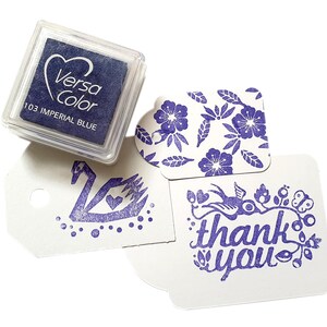 Pode incluir: Uma almofada de tinta azul com a etiqueta "VersaColor 103 Imperial Blue" com tr&ecirc;s imagens carimbadas: um cisne, um desenho floral e uma mensagem de "obrigado" com um p&aacute;ssaro e flores.