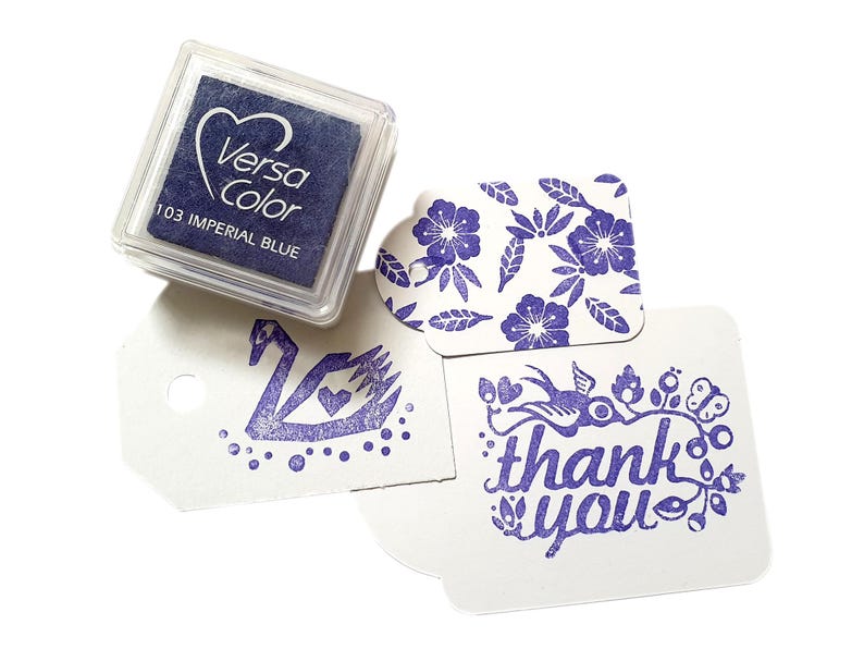 Pode incluir: Uma almofada de tinta azul com a etiqueta "VersaColor 103 Imperial Blue" com tr&ecirc;s imagens carimbadas: um cisne, um desenho floral e uma mensagem de "obrigado" com um p&aacute;ssaro e flores.