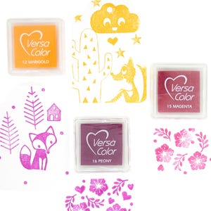 Pode incluir: Um conjunto de tr&ecirc;s almofadas de tinta para carimbar, com uma almofada amarela etiquetada "Versa Color 12 Marigold", uma almofada magenta etiquetada "Versa Color 15 Magenta" e uma almofada roxa etiquetada "Versa Color 16 Peony".