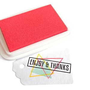 Pu&ograve; includere: Un tampone d'inchiostro rosso con una custodia in plastica bianca. Il tampone d'inchiostro viene utilizzato per timbrare la frase "ENJOY & THANKS" su un'etichetta bianca con un design geometrico colorato.