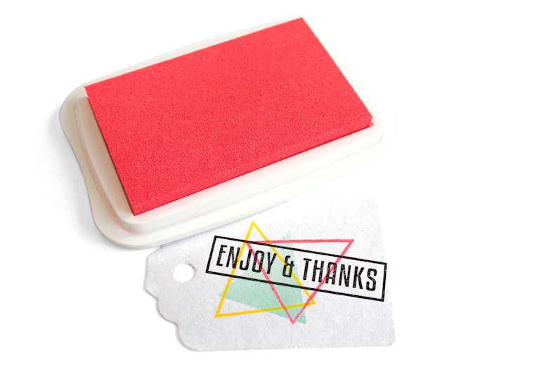 Pu&ograve; includere: Un tampone d'inchiostro rosso con una custodia in plastica bianca. Il tampone d'inchiostro viene utilizzato per timbrare la frase "ENJOY & THANKS" su un'etichetta bianca con un design geometrico colorato.