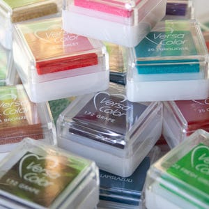 Pode incluir: Uma cole&ccedil;&atilde;o de pequenos recipientes de pl&aacute;stico transparente com almofadas de tinta coloridas dentro. Cada recipiente tem um r&oacute;tulo com o nome da marca "Versa Color" e um nome de cor, como "Sage", "Turquoise" e "Cardinal".
