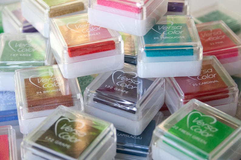 Pode incluir: Uma cole&ccedil;&atilde;o de pequenos recipientes de pl&aacute;stico transparente com almofadas de tinta coloridas dentro. Cada recipiente tem um r&oacute;tulo com o nome da marca "Versa Color" e um nome de cor, como "Sage", "Turquoise" e "Cardinal".