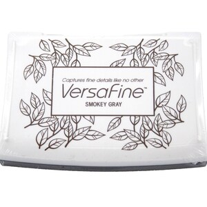 Puede incluir: Una almohadilla de tinta blanca con un borde negro y un dise&ntilde;o de hojas marrones. El texto "Captures fine details like no other VersaFine&trade; Smokey Gray" est&aacute; impreso en la almohadilla.