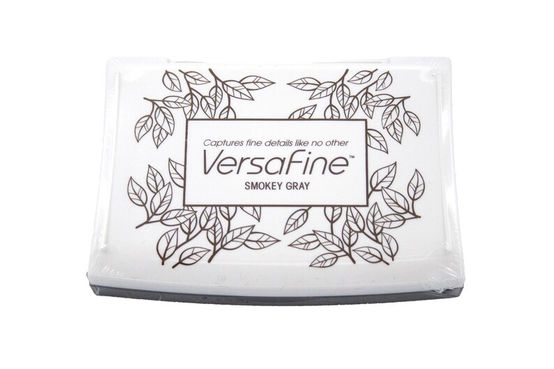 Puede incluir: Una almohadilla de tinta blanca con un borde negro y un dise&ntilde;o de hojas marrones. El texto "Captures fine details like no other VersaFine&trade; Smokey Gray" est&aacute; impreso en la almohadilla.