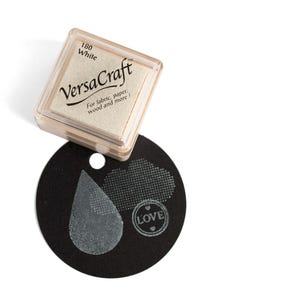 K&ouml;nnte beinhalten: Ein wei&szlig;es Stempelkissen mit der Aufschrift "VersaCraft", der Nummer "180" und dem Text "F&uuml;r Stoff, Papier, Holz und mehr!" Das Stempelkissen liegt auf einem schwarzen Kreis mit einem wei&szlig;en Stempelabdruck eines Tr&auml;nen tropfens, einer Wolke und dem Wort "LOVE" mit zwei Herzen.