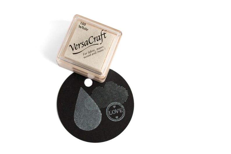 K&ouml;nnte beinhalten: Ein wei&szlig;es Stempelkissen mit der Aufschrift "VersaCraft", der Nummer "180" und dem Text "F&uuml;r Stoff, Papier, Holz und mehr!" Das Stempelkissen liegt auf einem schwarzen Kreis mit einem wei&szlig;en Stempelabdruck eines Tr&auml;nen tropfens, einer Wolke und dem Wort "LOVE" mit zwei Herzen.