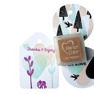 Pode incluir: Uma almofada de tinta marrom com o texto "VersaColor 182 Bisque". A almofada de tinta est&aacute; ao lado de uma etiqueta branca com o texto "Thanks & Enjoy" e um desenho de elefante roxo. H&aacute; tamb&eacute;m uma etiqueta branca com um p&aacute;ssaro preto e um desenho de gota preta.