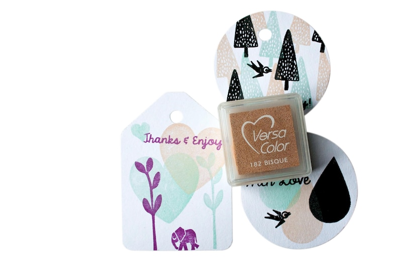 Pode incluir: Uma almofada de tinta marrom com o texto "VersaColor 182 Bisque". A almofada de tinta est&aacute; ao lado de uma etiqueta branca com o texto "Thanks & Enjoy" e um desenho de elefante roxo. H&aacute; tamb&eacute;m uma etiqueta branca com um p&aacute;ssaro preto e um desenho de gota preta.