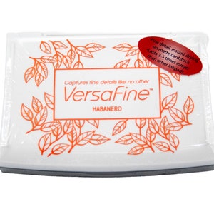 Puede incluir: Una almohadilla de tinta blanca con una etiqueta roja que dice "Fine detail, instant drying on matte cardstock. Lasts 3-5 times longer than other inkpads." La almohadilla de tinta est&aacute; etiquetada como "VersaFine" y "HABANERO" en texto naranja.