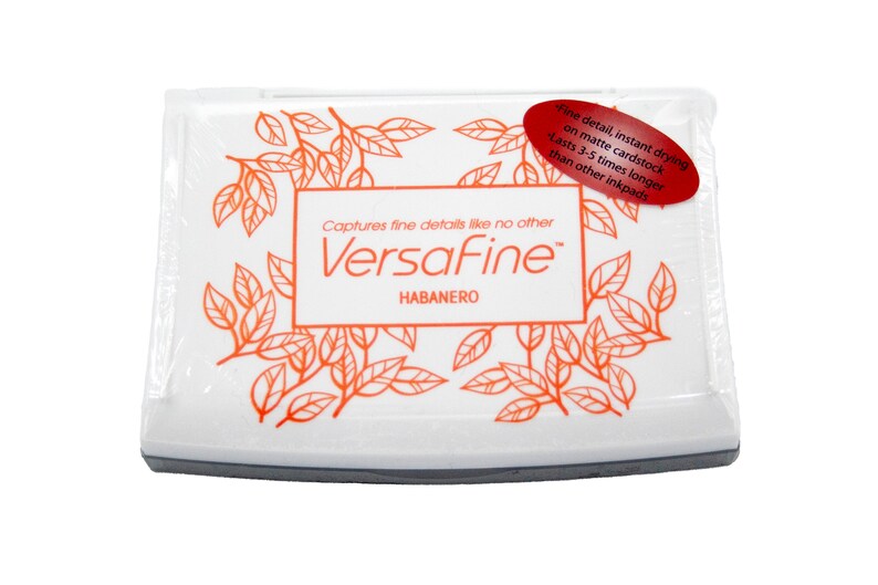 Puede incluir: Una almohadilla de tinta blanca con una etiqueta roja que dice "Fine detail, instant drying on matte cardstock. Lasts 3-5 times longer than other inkpads." La almohadilla de tinta est&aacute; etiquetada como "VersaFine" y "HABANERO" en texto naranja.