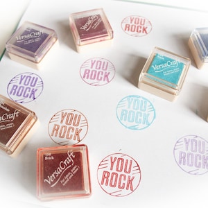 K&ouml;nnte beinhalten: Eine Sammlung von neun Stempelfarben mit dem aufgedruckten Text "You Rock". Die Stempelfarben sind in verschiedenen Farben, darunter Rot, Blau, Gr&uuml;n und Lila. Die Stempelfarben sind in einem Rastermuster auf einer wei&szlig;en Oberfl&auml;che angeordnet.